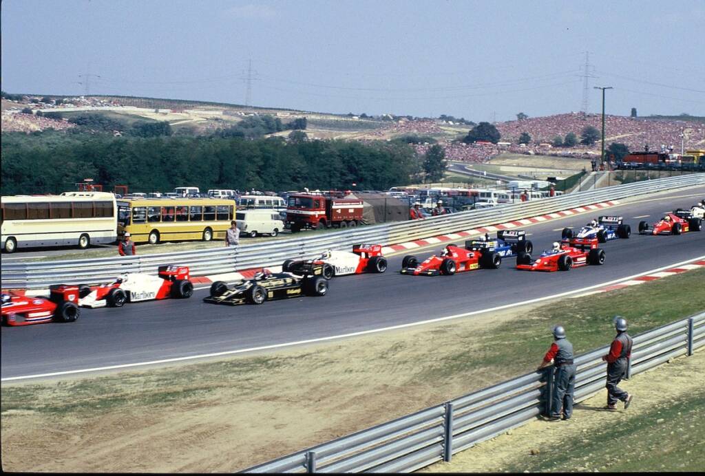 1980s Formula 1 cars taking a corner at the Hungaroring with marshals observing behind the barrier.