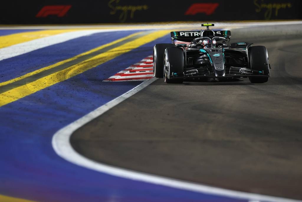 A black and teal Mercedes AMG Petronas F1 car speeding around a bend on the Marina Bay Street Circuit during the Singapore Grand Prix night race, with yellow and blue curbs in the foreground.