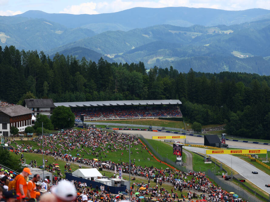Crowds fill the hillsides and grandstands at the Red Bull Ring as Formula 1 cars race past during the Austria Grand Prix, with forested mountains in the background.