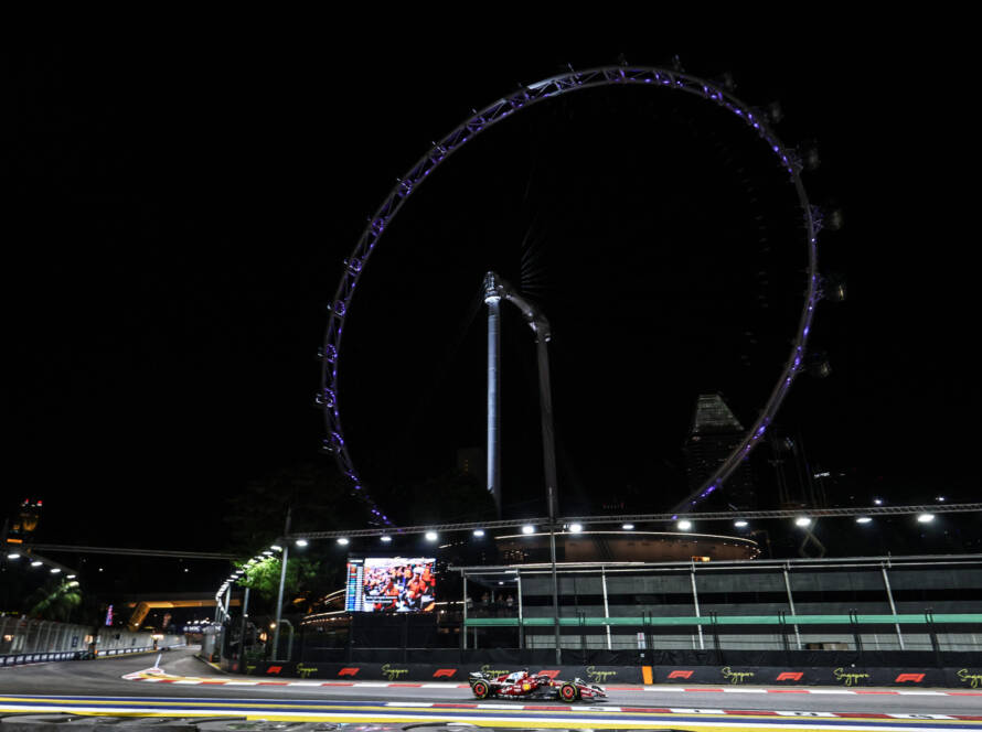 A Formula 1 car driving along the illuminated Marina Bay Street Circuit at night in Singapore with the Singapore Flyer ferris wheel lit up in the background.